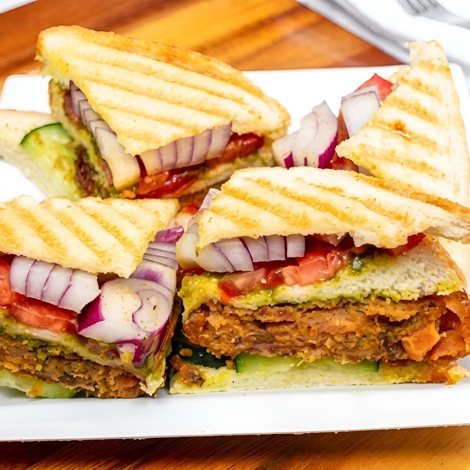 Grilled sandwich triangles layered with crispy filling, tomatoes, cucumbers, red onions, and green chutney on a white plate.